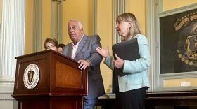 A man in a gray suit stands at a wooden lectern. Next to him, a woman in a blue jacket gestures while holding a binder.