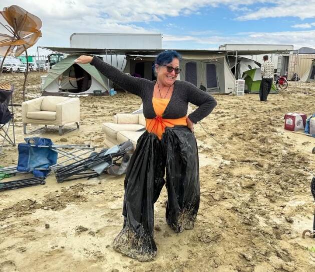A woman models garbage bag pants as she stands in the rain-soaked and muddy playa at Burning Man with chairs and tents behind her.