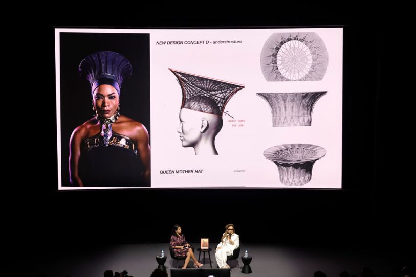 Academy Museum Of Motion Pictures Hosts The Art Of Ruth E. Carter: Book Signing And Conversation. In this photograph with E. Carter and another woman are sitting on a stage, overhead there is a large projection of one of Carter's costume designs for "Black Panther." It is a series of illustrations of how she made the headdress for Queen Ramonda, played by Angela Basset.