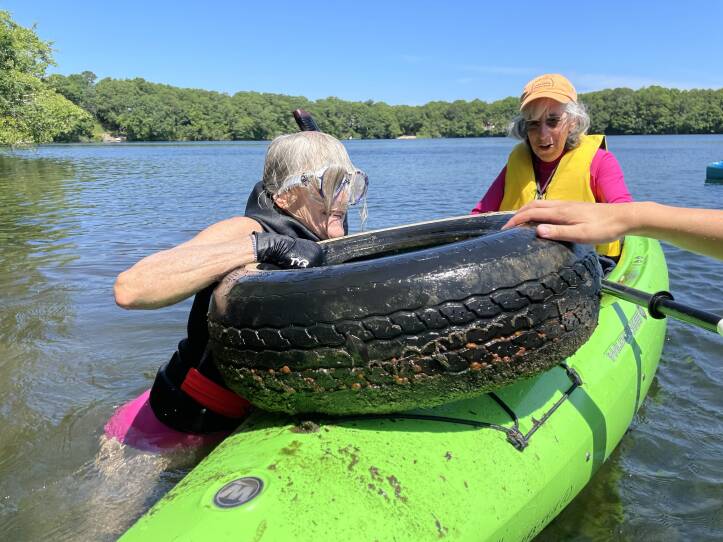 An older woman in a swimsuit and goggles lifts a tire onto the front of a green kayak that another older woman is paddling.