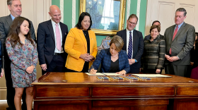 With several people behind her, a white woman with short brown hair sits at a wooden desk signing papers.