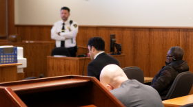 A young man in a suit and an older man in a camo winter jacket sit in a courtroom. A bailiff is seen in the background.
