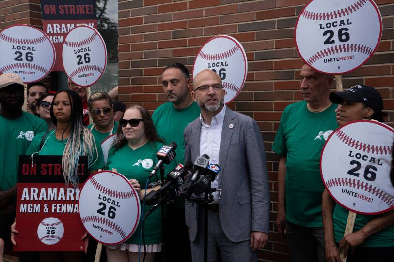A man in a gray suit and glasses speaks into several news microphones, while people in green t-shirts cluster around him holding signs of large baseballs bearing the words "Local 26 UNITEHERE!"