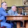 Two men in suits sit at a wooden table, surrounded by bookshelves.