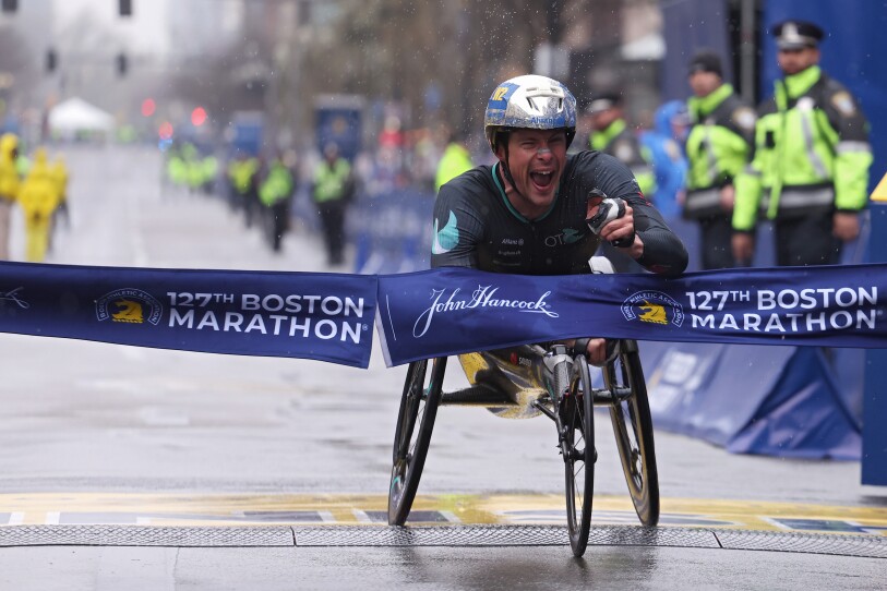 A man in a racing wheelchair breaks through the Boston Marathon tape at the finish line. He joyfully yells and waves his hand.