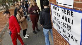People line up outside a brick building with a paper sign that says vote here 7AM-8PM in English, Spanish, Chinese and Vietnamese.