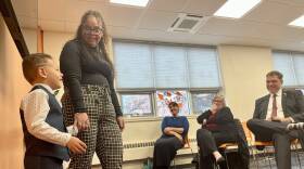 A young boy in a vest and bow-tie holds hands with his teacher as he addresses a classroom full of adults.