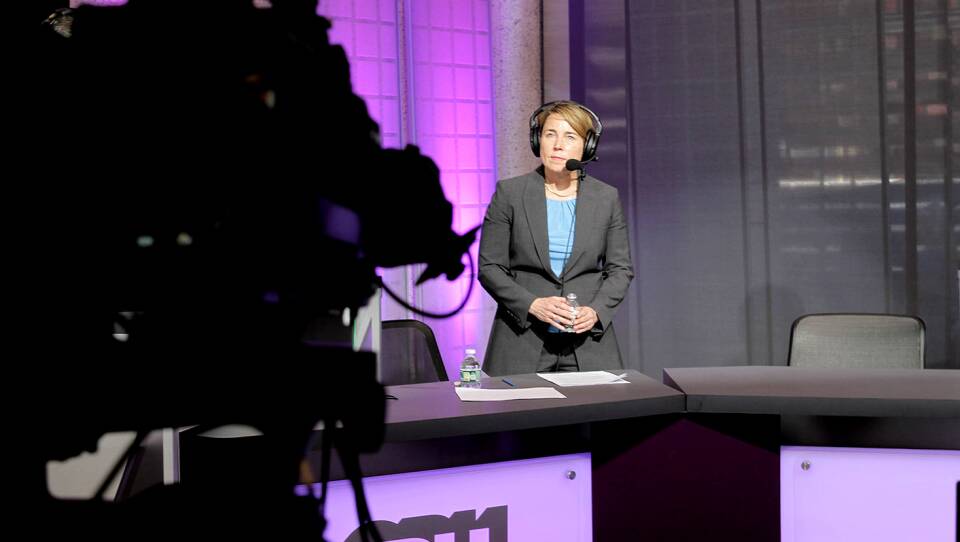 The governor stands on GBH's purple set in a radio headset and blazer, listening intently. She's facing a camera that stands in the foreground, obscured in the shadows.