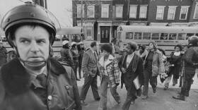 A police officer looks concerned as about a dozen students and other officers walk near school buses behind him.