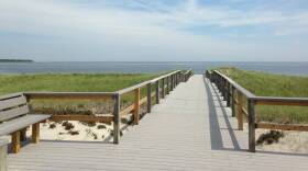 A raised wooden boardwalk leads out, over marsh grass and sand, toward a wide body of water.