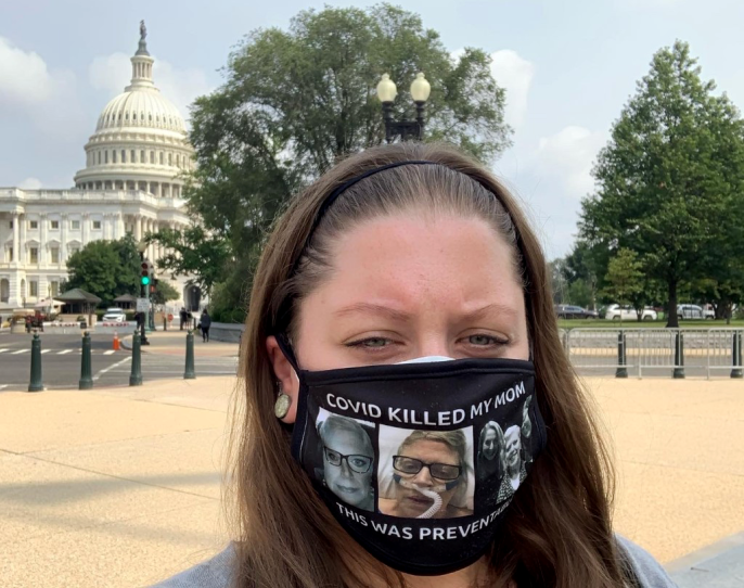 A white woman with long brown hair stands in Washington, D.C., with the Capitol building in the background wearing a mask that says "COVID killed my Mom, this was preventable" with three photos of her mother on it.