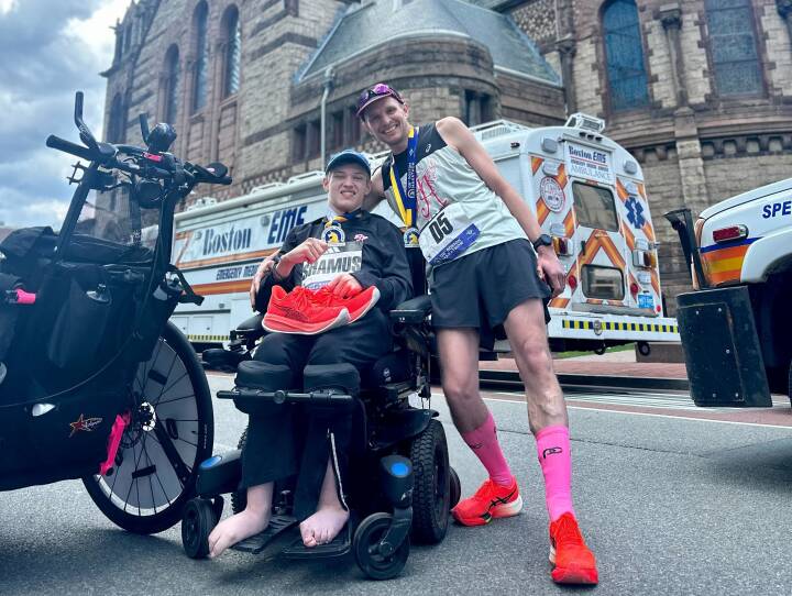 A father wraps one arm around his son in a wheelchair. Both are smiling and wearing race bibs affixed to their shirts.
