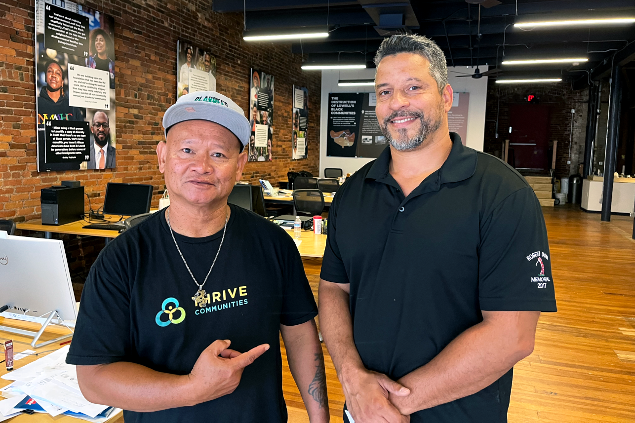 Two men stand in an open office, posing for the camera.