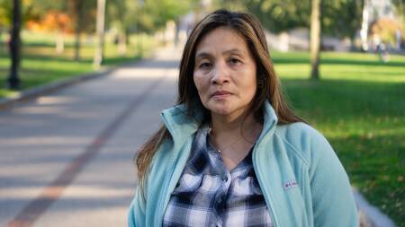 A woman looks calm but upset as she stands in a park during autumn, with some leaves changing colors behind her.