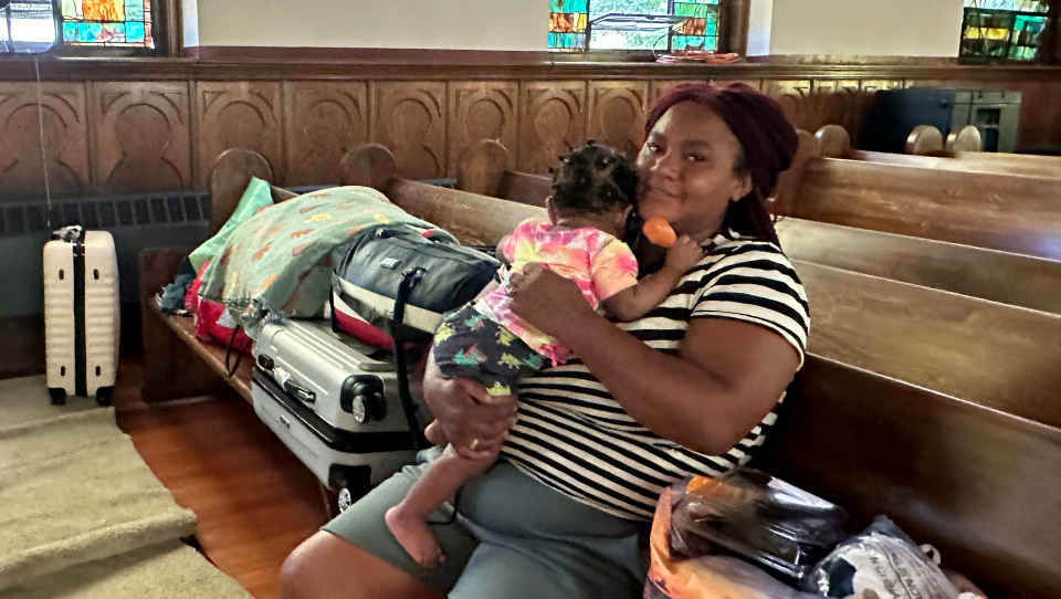 A woman sits in the front pew of a church while holding a young child. Bags and luggage cover the pew around her.