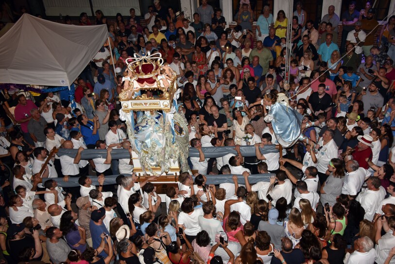 A girl held up by a rope and harness, wearing a light blue cape and angel wings, floats over a large crowd of people near a statue of the Virgin Mary.