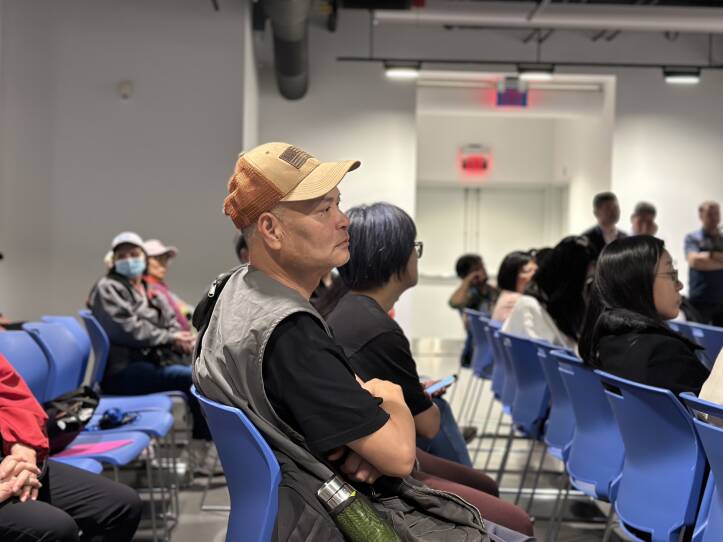 A man in an orange cap and grey vest sits with his arms crossed at a meeting.