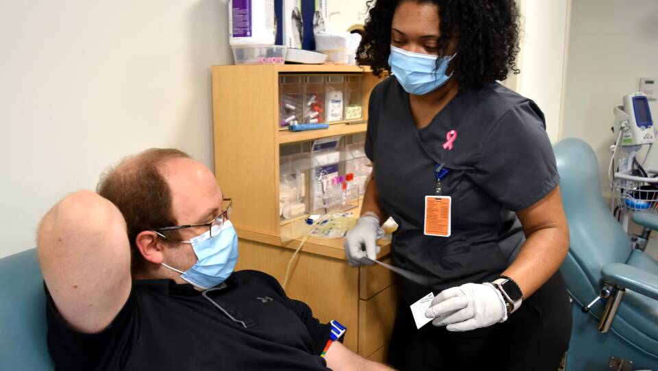A man lying in a blood donation chair extends one arm out in front of him, the other behind his head. The phlebotomist holds materials to prep him for donating blood in her hands as she moves toward him.
