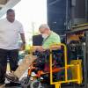 A man in a wheelchair sits on the lift on the back of a large bus. An operator stands next to him