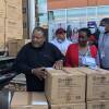Westnet CEO Gordon Thompson, wearing a pullover sweatshirt, stands beside a stack of cardboard crates, which he is preparing to help load onto a heavy truck, which can be seen in the background loaded with similar boxes. Beside him is Frederica Williams president and CEO of the Whittier Street Health Center, wearing a red sweater over a black and white patterned dress, holding a box of medical face masks.