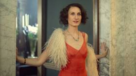 A dark haired woman in an orange 1930s costume stands in a fancy doorway