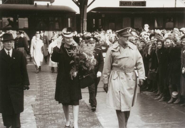Crown Princess Martha and King Olave arrive at Trondheim, Norway. Crowds cheer them on. Martha wears a black dress, a white hat with black feathers, and carries a large bouquet of wildflowers. Olav wears a trench coat and military cap.