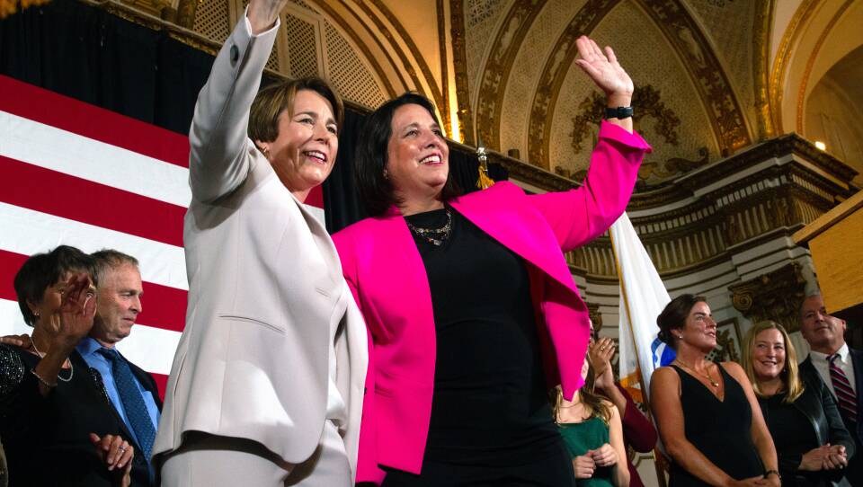 Two women wave and smile out at a crowd, one in a white pantsuit and one in a bright pink blazer. Others gather behind them in support in the background.