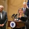 A woman stands at a podium branded with the Massachusetts state seal and speaks into a microphone, folders and a surgical mask in her hands