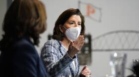 A woman wearing a blazer and respiratory mask gestures to a group, seated at a table during a meeting