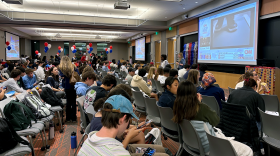Students with backpacks sit in plastic chairs in what appears to be a lecture hall. CNN feeds are projected onto screens. Red white and blue decorations like balloons are around the room.