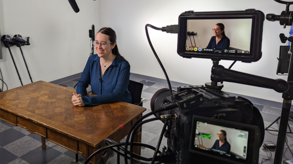 Claire Houston sits at a desk in a video recording studio. Two video screens show her smiling.
