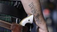 A close-up photo shows a white-handled gun in a hip holster next to a person's arm that has a tattoo reading "we the people"