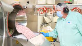 A worker in protective clothing and headphones holds the side of a metal chute as pink pills cascade into it.