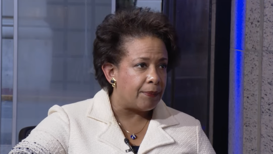 Loretta Lynch has a slight smile as she listens to a reporter, who is just out of the frame, in a studio space.