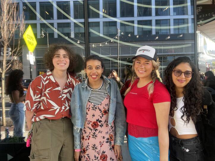 Four friends stand next to each other in front of a building. It is a sunny day and appears to be warm as everyone is wearing lightweight clothing.