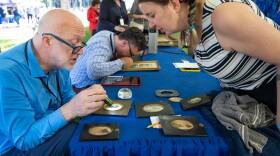 A man seated at a table inspects the details of a small framed object while its owner leans over, observing.