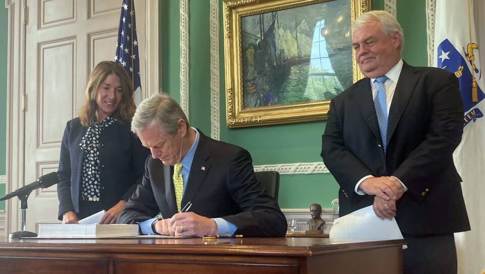 Gov. Charlie Baker, seated at a desk, signs papers as Lt. Gov. Karyn Polito and Administration and Finance Secretary Michael Heffernan stand behind him.