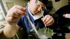 A man wearing scrubs uses a long tool to delicately remove a vial from a cylindrical container in a laboratory.