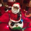 A small figurine of a Black Santa Claus sits on a table surrounded by multicolored string lights and other figurines.
