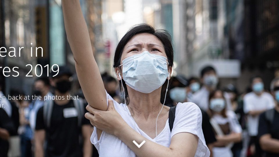A women in a surgical mask, eyes looking up, raises her arm. A crowd of masked people stands behind her. Header image for 'Year in Pictures'