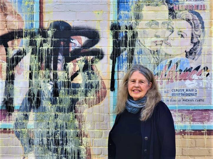 A woman wearing a scarf smiles at the camera and stands in front of a painted brick wall, covered in faded graphics and graffiti