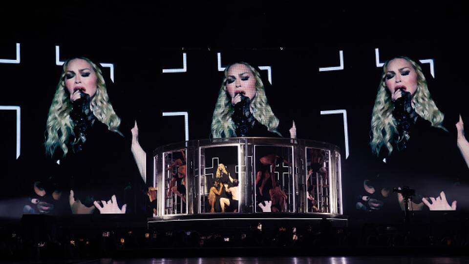 Madonna performs on stage. She is in surrounded by panels with dancers in provocative clothing around her. Behind them are three large screens showing Madonna singing into a microphone.