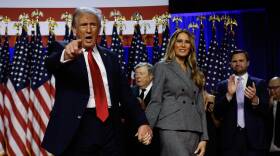 Donald Trump holds hands with Melania on stage, pointing to the crowd. Vice President Elect JD Vance claps in the background.