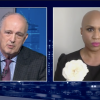 Split screen with jim braude, wearing a suit and a purple tie who is mid-speech, and ayanna pressley on the right, wearing a black top with a large white flower pinned to it, with the "greater boston" logo in the bottom left corner