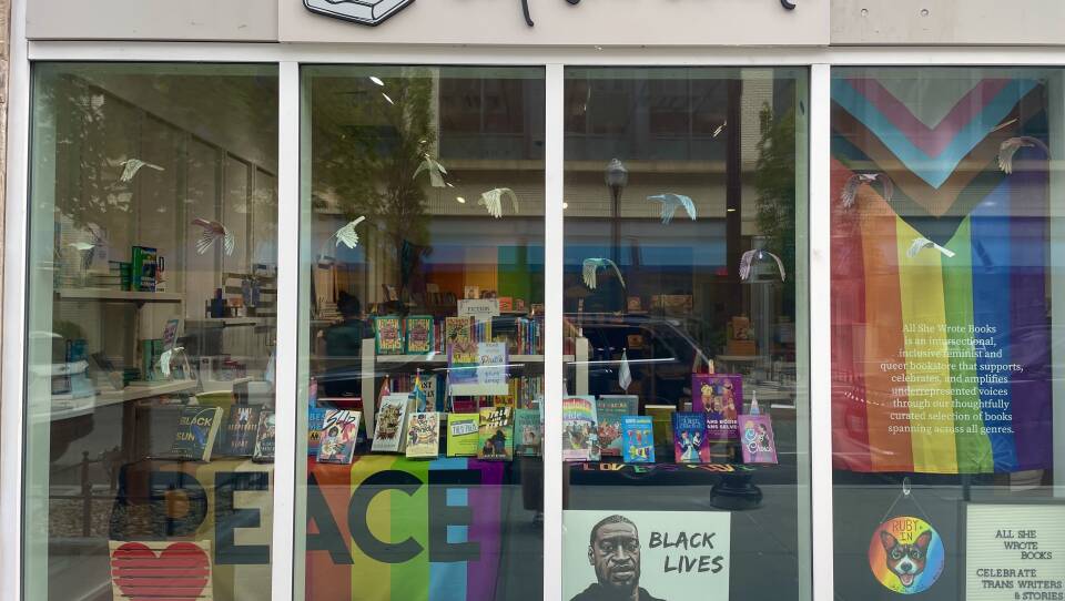 This is a photograph of a storefront of a book store. Hanging in the window is a multi colored striped Progress Pride flag, there is also a Pride Flag. in the storefront window is a table, which has  a display of books propped on it and a Pride flag draped over it. Propped up in the window sill is a Black Lives Matter poster with the face of George Floyd , a painting of a red heart and a sign in the window, which says "All She Wrote Books. Celebrate Trans Writers & Stories Every Day"