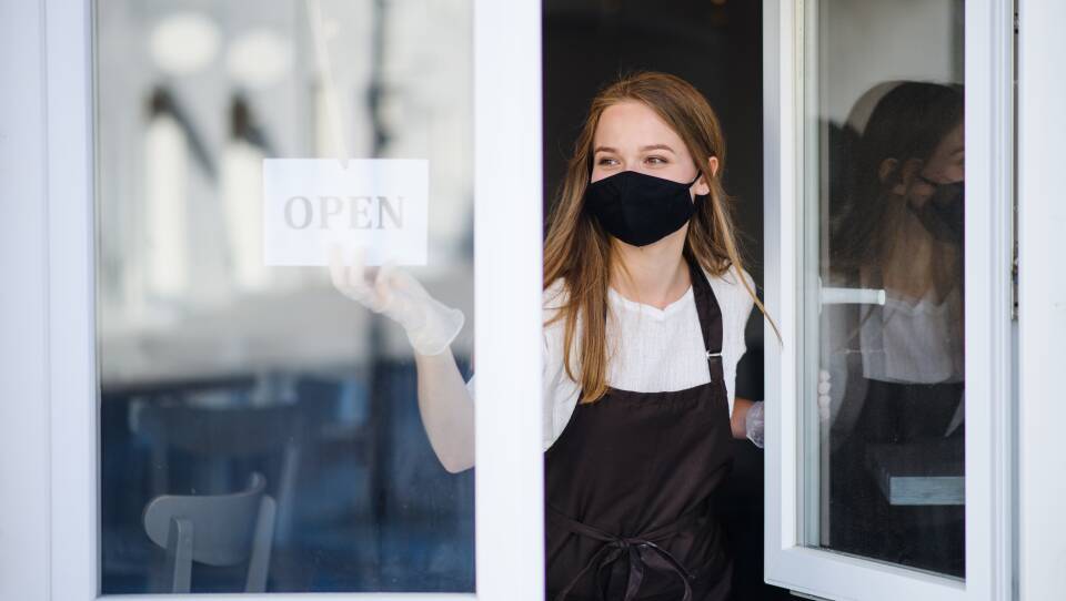 A woman wearing a face mask and gloves opens a shop window.