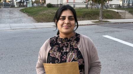 A woman wearing a sweater stands on the sidewalk of a residential neighborhood.