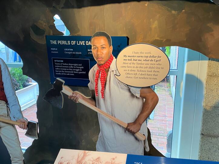 A museum exhibit includes a modern life-sized photo of a black man dressed as an enslaved worker carrying an axe