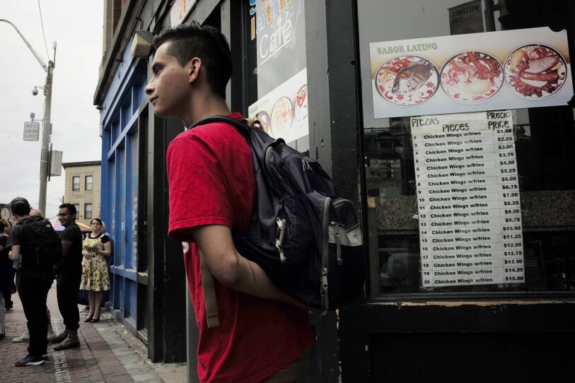 A teenager wearing a backpack stands next to a restaurant. Other people are on the sidewalk around the corner.