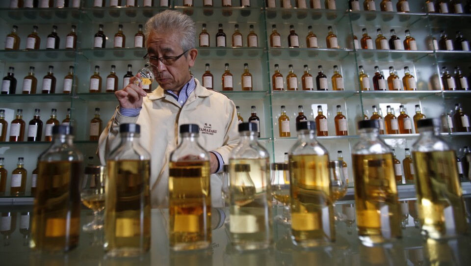 A man in a white coat holds a glass containing whisky, swirling the crystalline amber spirit against the light.